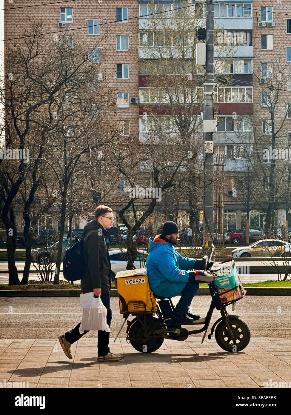 Zustellkurier unterwegs, vorbei an einem Fußgänger auf einer Stadtstraße, der den modernen urbanen Lebensstil und die Lieferung von Speisen unterstreicht. - Smartphone-aufgenommenes Stockfoto
