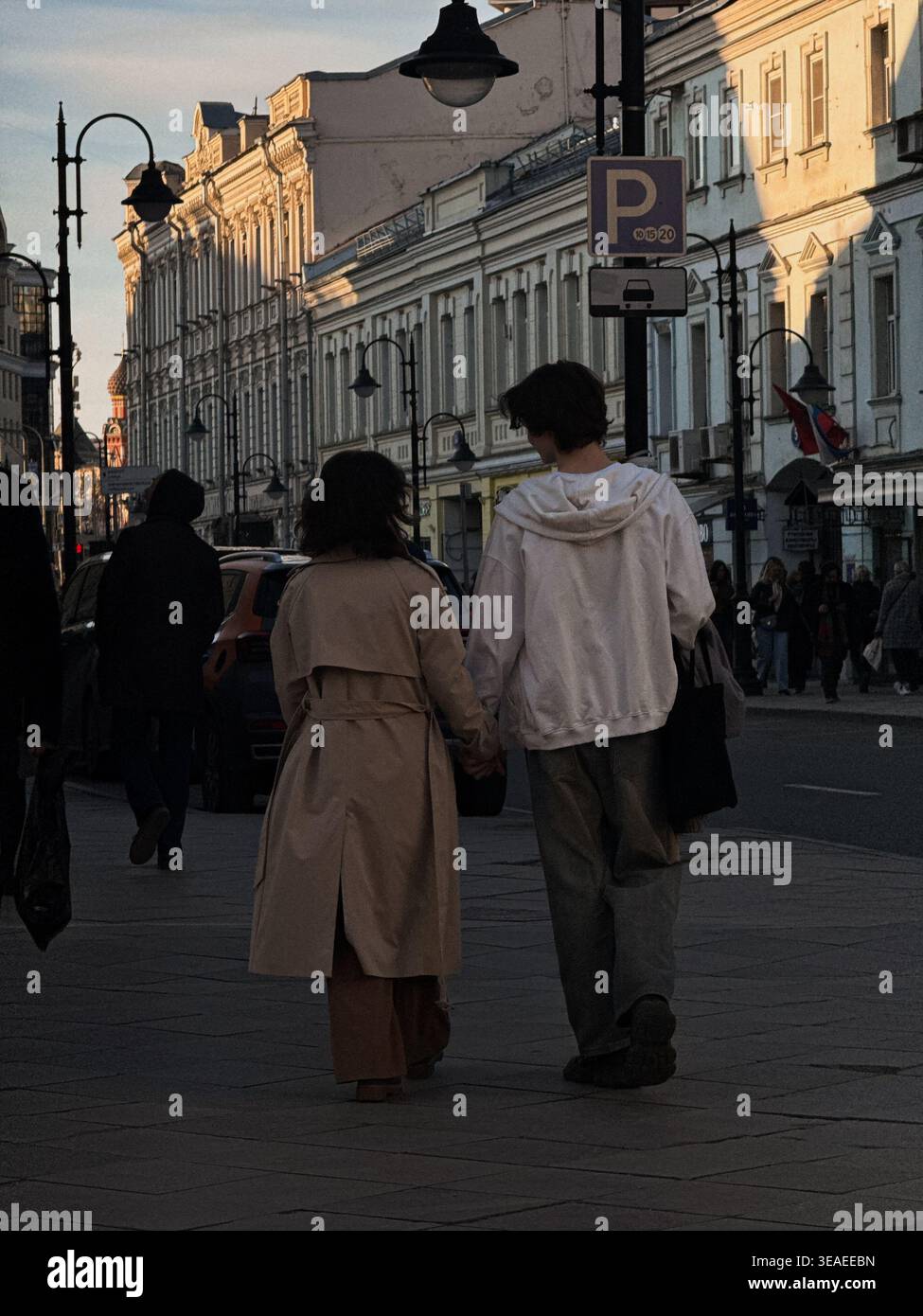 Romantisches junges Paar, das Hände hält und zusammen in der Stadt bei einem goldenen Sonnenuntergang spaziert, mit Silhouetten von städtischen Gebäuden - Smartphone-aufgenommenes Stockfoto