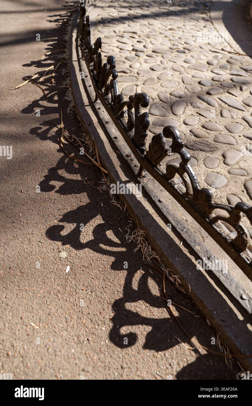Niedriger geschmiedeter Eisenzaun neben einem Kieselweg in einem Stadtgarten unter hellem Sonnenlicht. Stockfoto