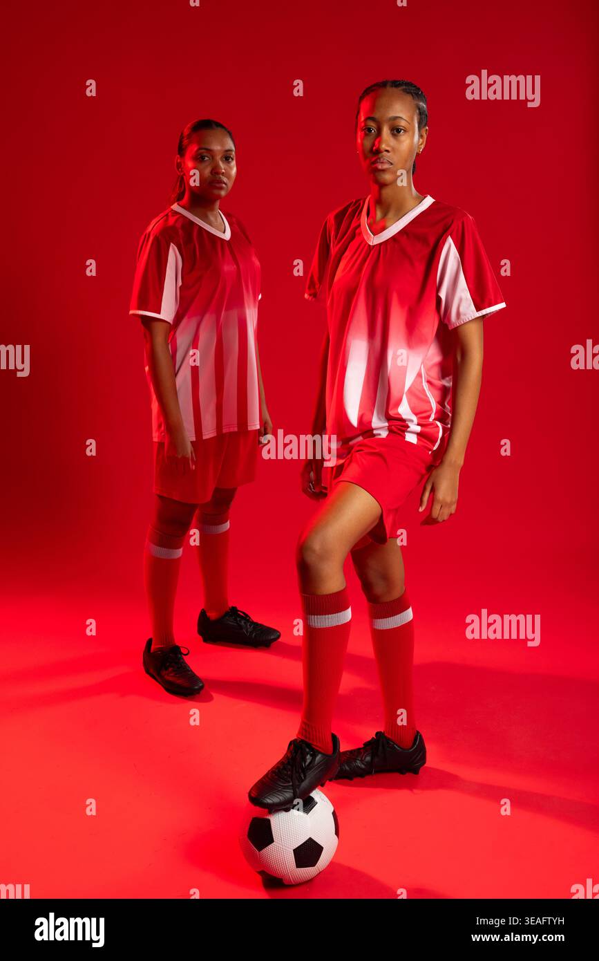 Weibliche Mannschaftskameraden posieren im roten Studio, setzen den Fuß auf einen schwarz-weißen Fußball und tragen Uniformen Stockfoto