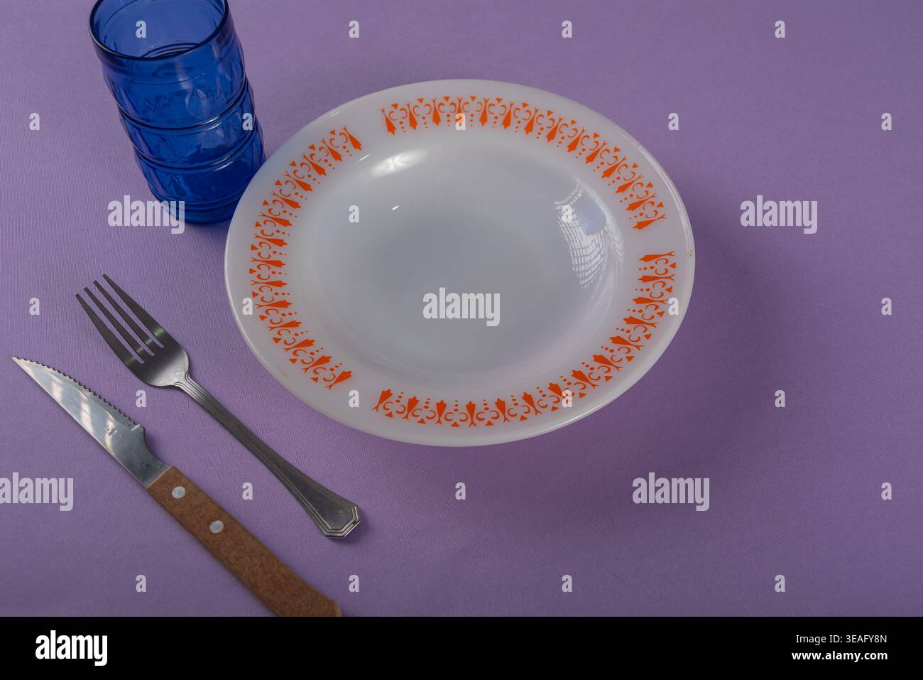 Stillleben zeigt eine einfache Tischeinstellung auf violettem Hintergrund. Mit Geschirr und sanfter Beleuchtung. Weiß-, Orange- und Blautöne. Kommerzielle Verwendung für Lebensmittel Stockfoto