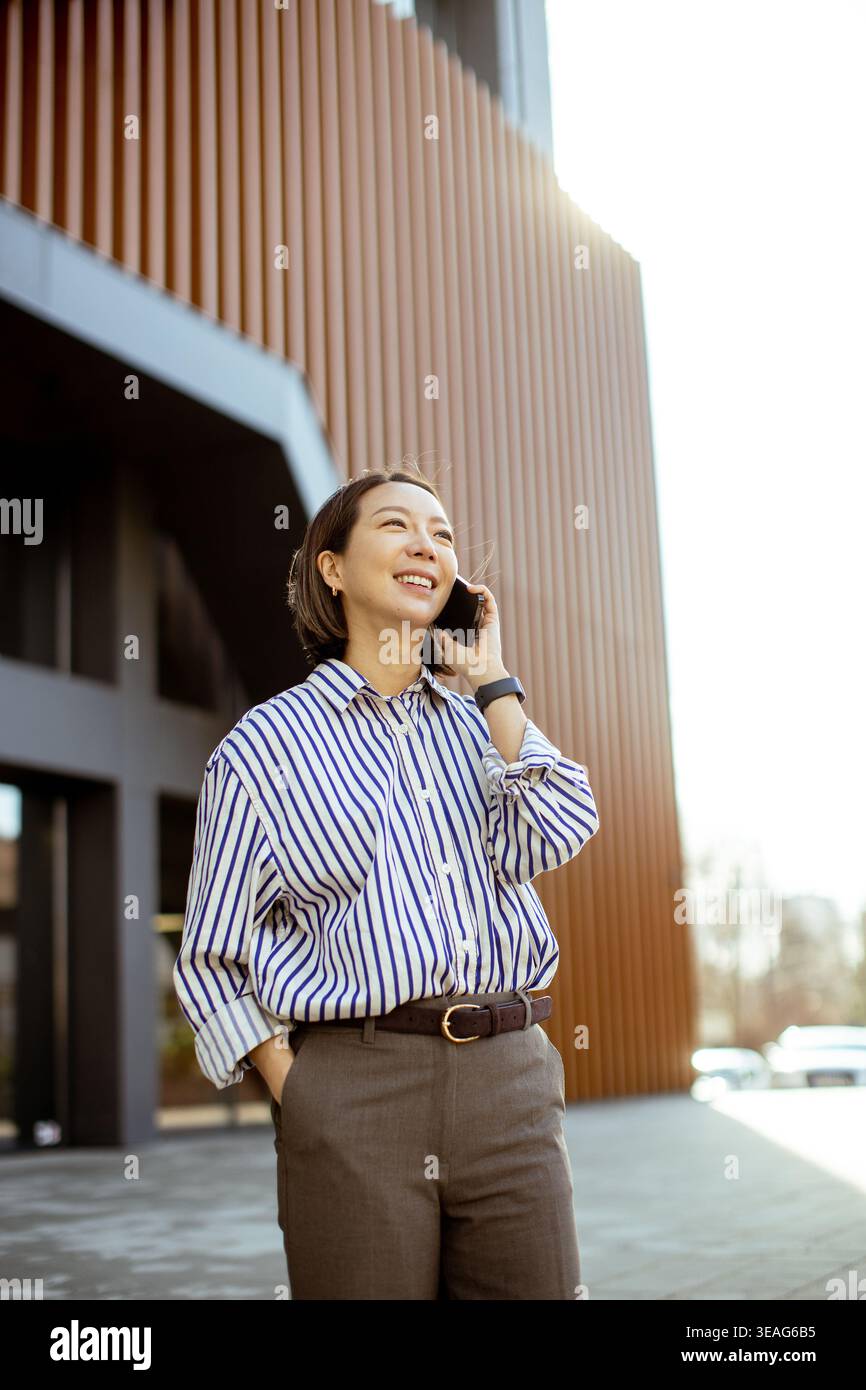 Eine fröhliche Koreanerin steht vor einem modernen Gebäude und unterhält sich mit einem hellen Lächeln am Telefon. Die Sonne scheint warm oben. Stockfoto