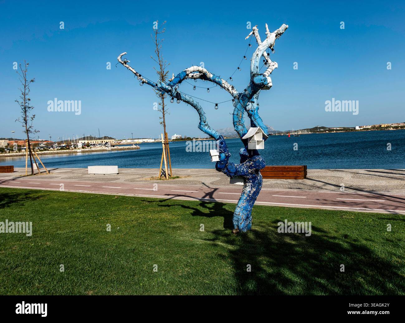 Eine blau bemalte Baumskulptur mit Vogelhäusern und Lichtern steht auf einem Uferweg in Olbia, Sardinien, vor einem klaren blauen Himmel und Hafen. Stockfoto