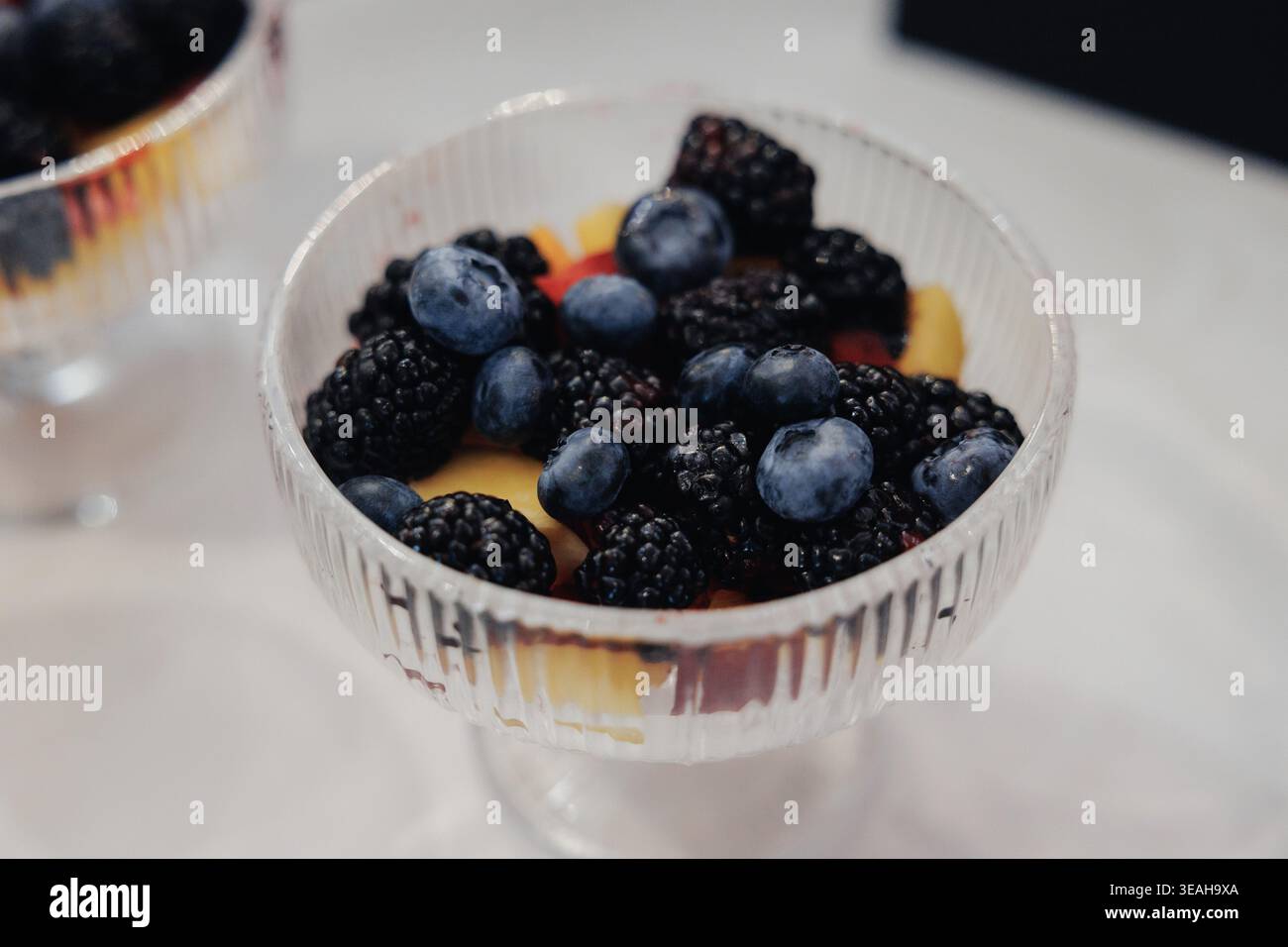 Frische Heidelbeeren Brombeeren In Kristallschale, Pfirsichscheiben Für Warmen Kontrast, Weiches Natürliches Licht, Geringe Schärfentiefe, Minimalistischer Weißer Tisch Stockfoto