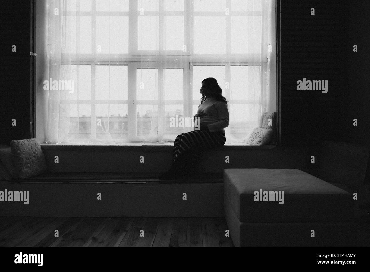 Silhouettenfrau, Die Am Fenster Sitzt, Weiches, Diffuses Licht Und Tiefe Schatten, Stadtbild Über Glas Hinaus Verschwommen, Minimal Interior Mit Breiter Fensterbank Stockfoto