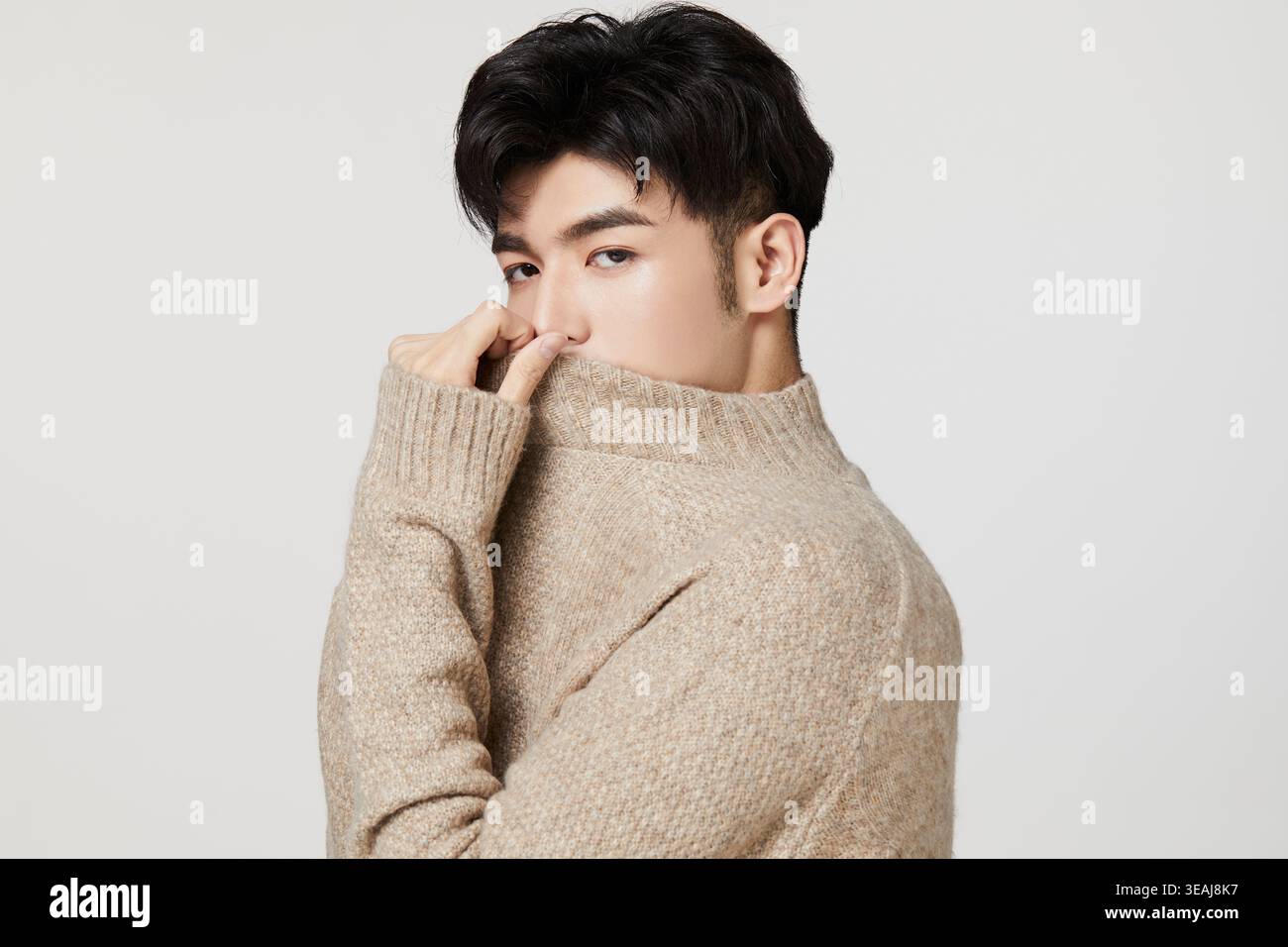 Ein junger asiatischer Mann im Alter von 20 zieht seinen Pullover über den Mund und blickt mit einem lässigen Ausdruck von der Kamera weg. Stockfoto