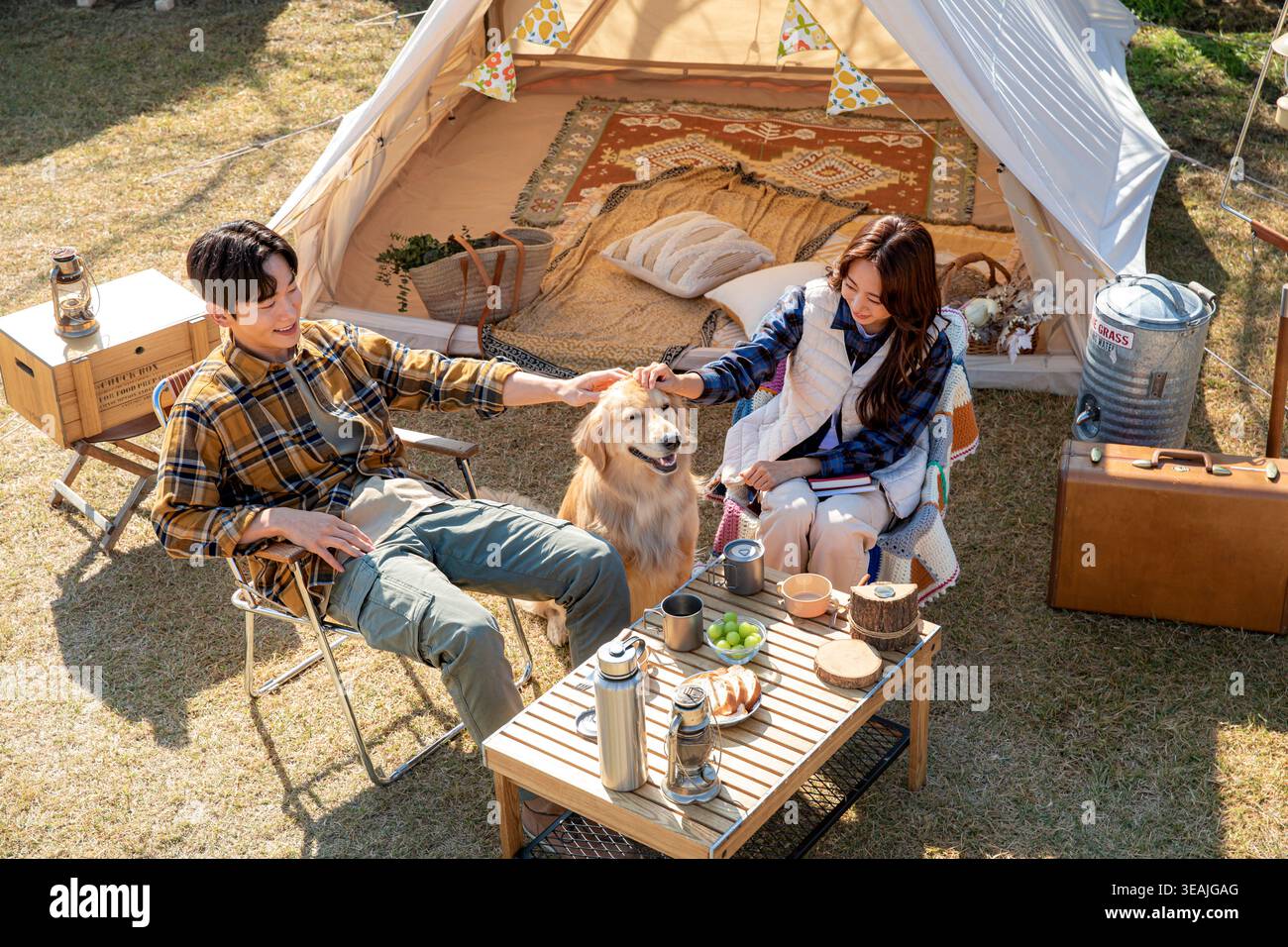 Ein Mann und eine Frau sitzen auf einem Campingplatz und streicheln einen goldenen Retriever, während sie draußen essen. Stockfoto