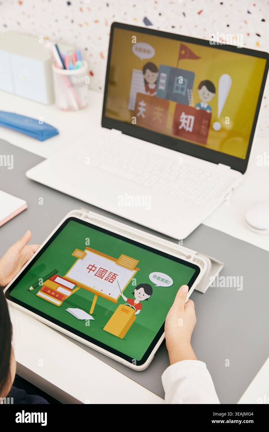 Eine Person verwendet ein Tablet, um einen Bildschirm mit chinesischen Schriftzeichen und eine Illustration einer Frau im Zeichentrickstil anzuzeigen, die auf einer grünen Tafel unterrichtet. Stockfoto