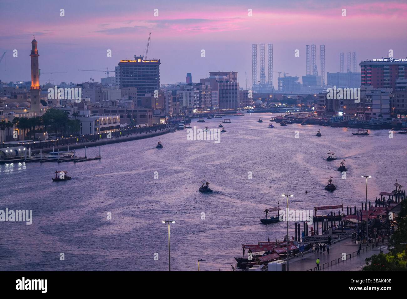 Blick aus der Vogelperspektive auf traditionelle hölzerne Abra-Boote, die das ruhige Wasser des Dubai Creek während einer violetten Dämmerung mit Stadtgebäuden und einem Moschee-Minarett überqueren Stockfoto