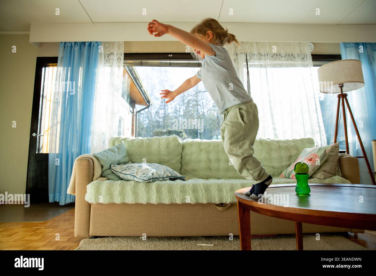 Niedrigwinkelaufnahme eines fröhlichen kleinen Kindes mit langen Haaren, das vom Sofa springt, voller Energie und verspieltem Lachen während eines hellen morgens zu Hause Stockfoto