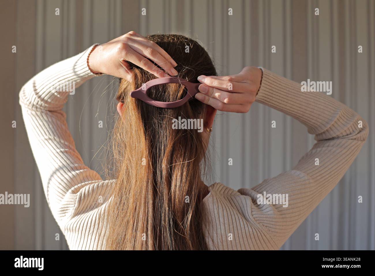 Frau, Die Langes Haar Mit Klauenklammer Stylt. Rückansicht einer Frau, die ihr langes braunes Haar mit einem rosafarbenen Krallenclip in warmem, natürlichem Innenlicht fixiert. Stockfoto