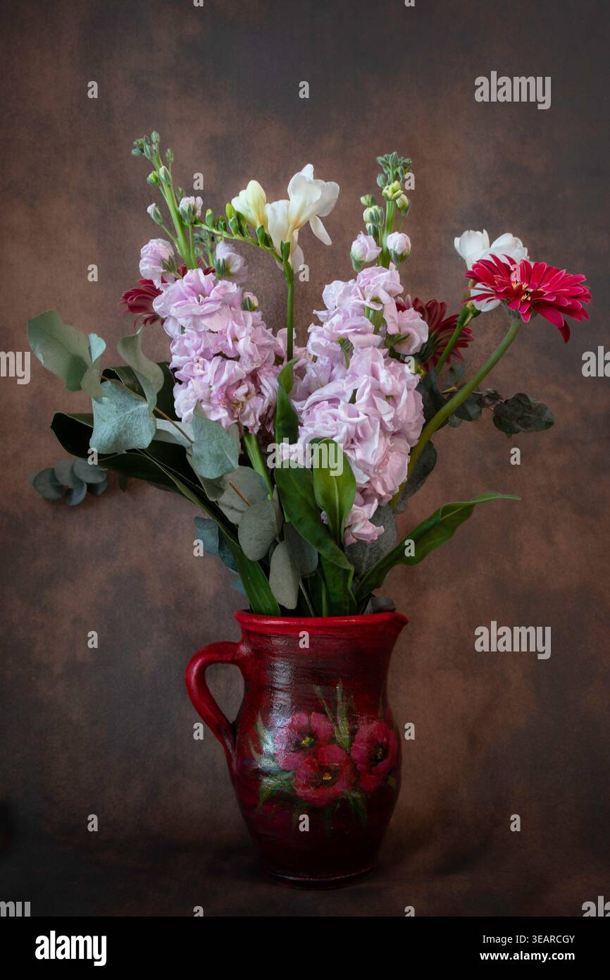 Vertikales Stillleben eines Blumenstraußes mit rosa Brühe, weißer Freesie und roter Gerbera in einem handbemalten roten Keramikkrug vor dunklem Hintergrund. Stockfoto
