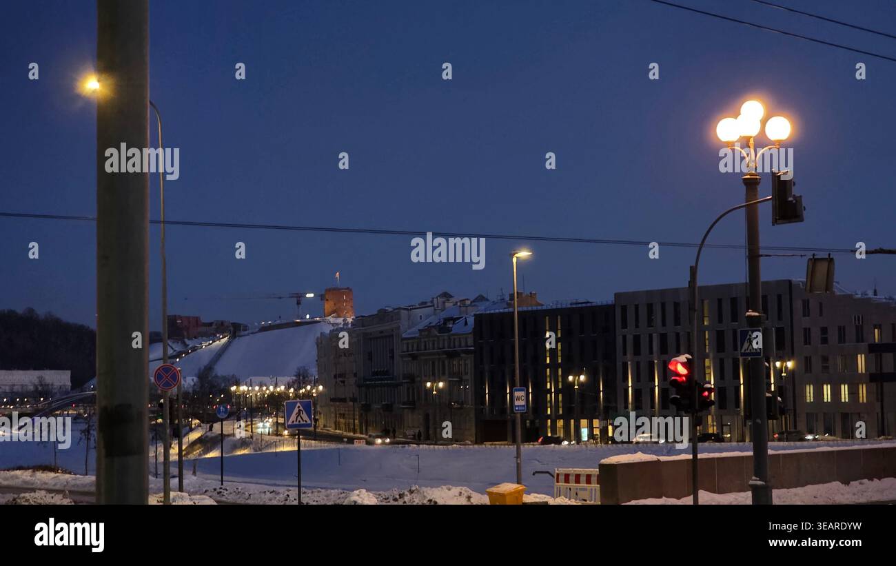 Vilnius Stadtzentrum zur blauen Stunde im Winter mit dem Turm der Burg Gediminas auf einem schneebedeckten Hügel, Straßenampeln und Ampeln im Vordergrund, Vilnius. - Smartphone-aufgenommenes Stockfoto