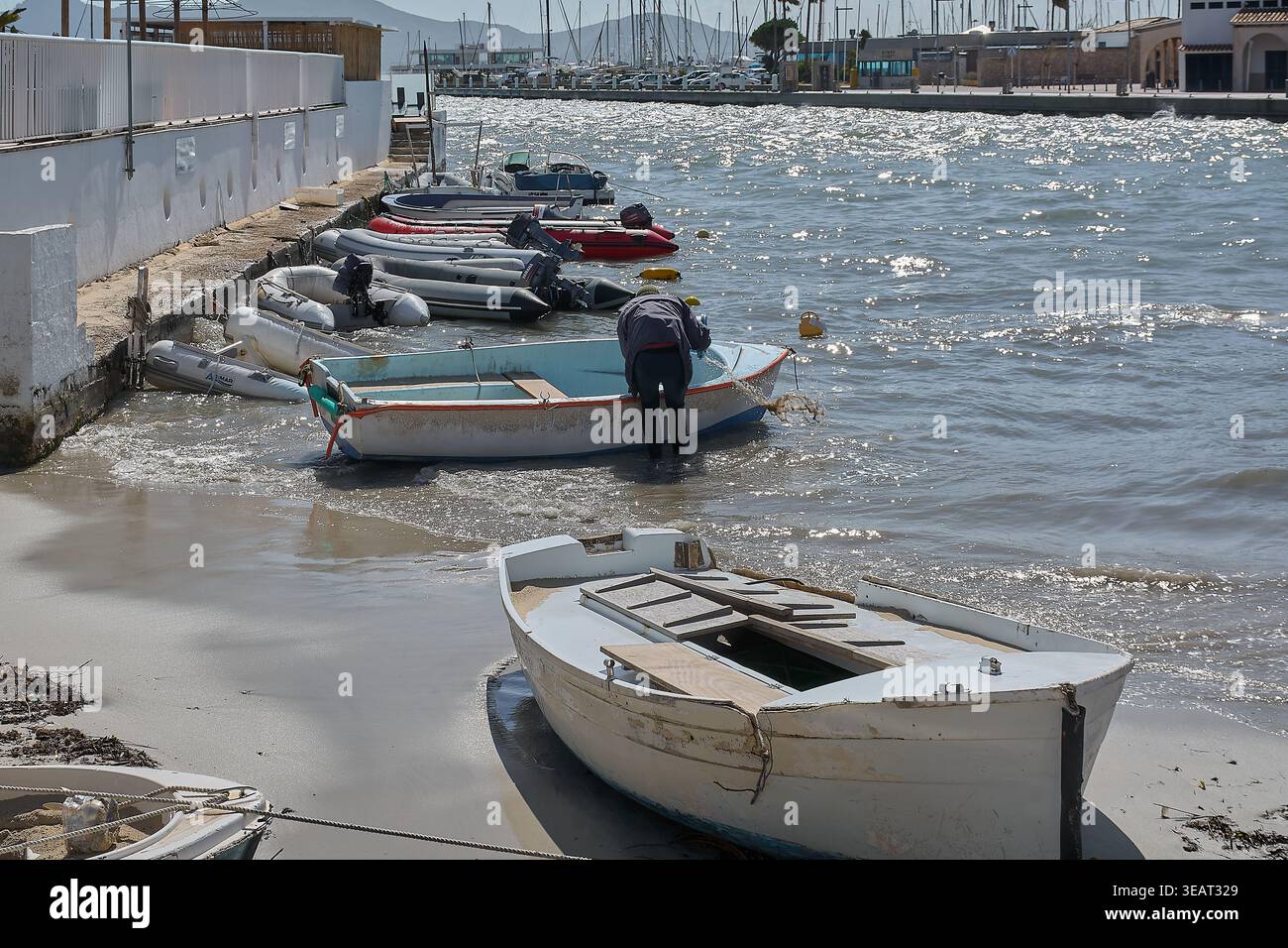 Mallorca Spanien - 23. April 2026: Ein einheimischer Fischer in einer dunklen Jacke und einem Taucheranzug zieht ein Netz in ein kleines weißes und blaues Boot am Wasserrand in a b Stockfoto