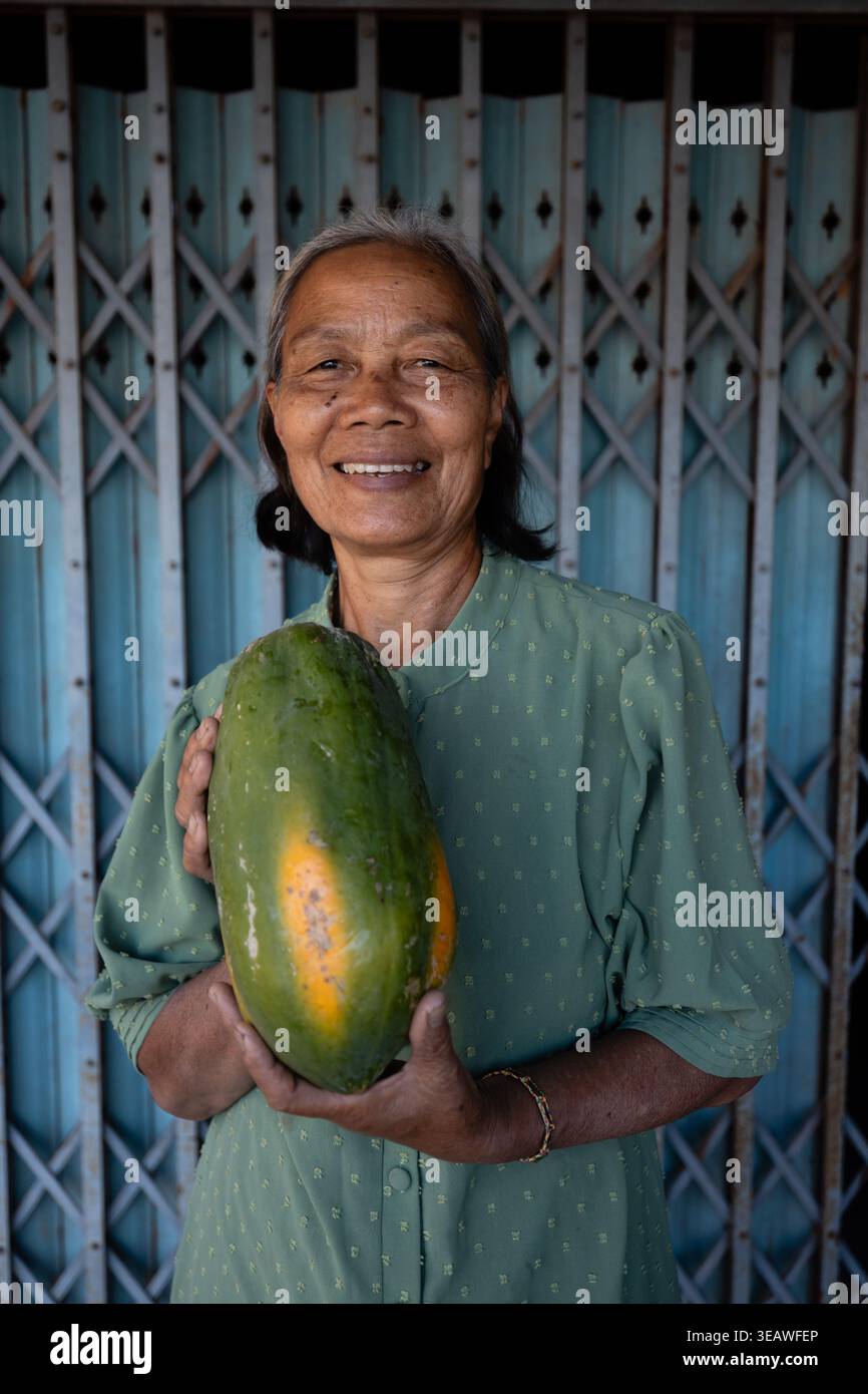 Loei, Thailand - 18. Dezember 2025: Eine lächelnde thailändische Straßenverkäuferin mit reifer Papaya-Frucht in der Nähe der Grenze zu Thailand und Laos. Ban Muang Stockfoto