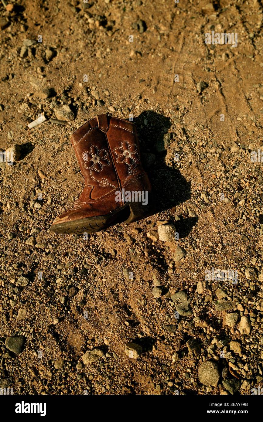 Ein einziger Cowboystiefel für Kinder, der auf trockenem Boden in einer Wüstenlandschaft liegt, deutet auf Verlassenheit oder ländliche Americana-Themen hin. Stockfoto