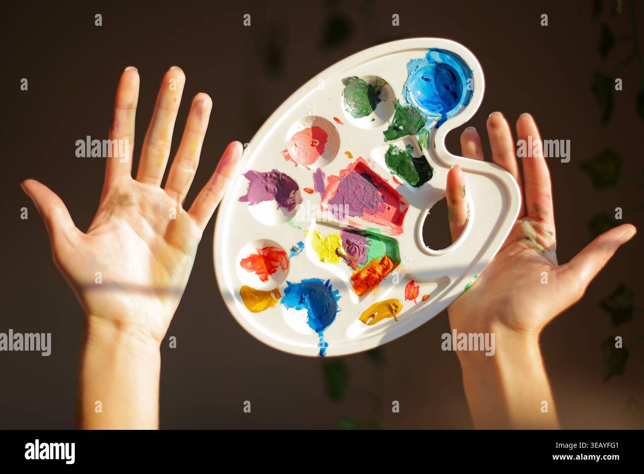 Malpalette Und Farbenfrohe Künstlerhände. Die Hände sind mit hellen Farben bedeckt und enthalten eine Künstlerpalette, die Kreativität, Farbmischungen und verspielt zeigt Stockfoto