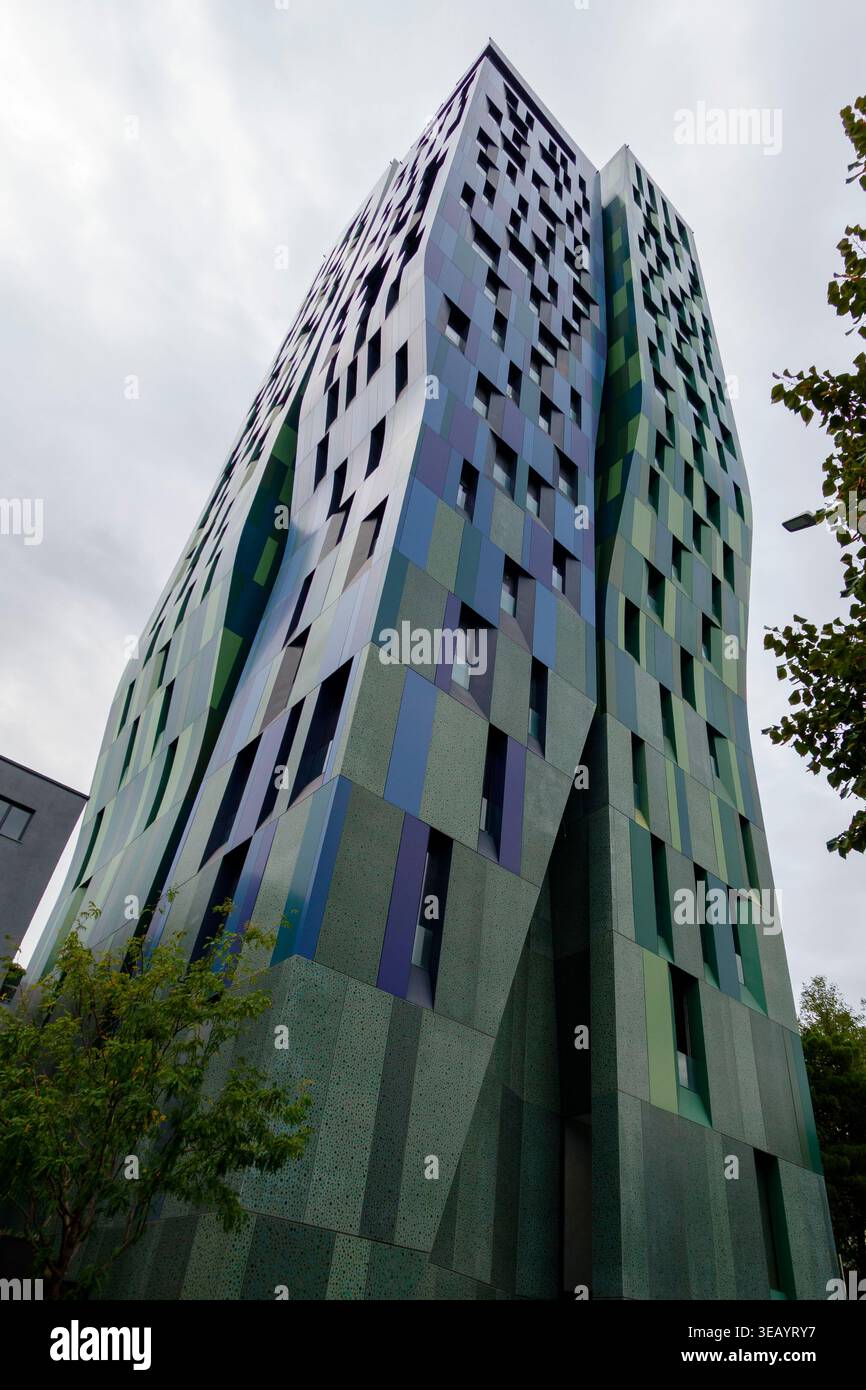 Moderner welliger Wolkenkratzer mit bunten grünen blauen und violetten Fassadenpaneelen unter bewölktem Himmel in einer städtischen Umgebung Stockfoto