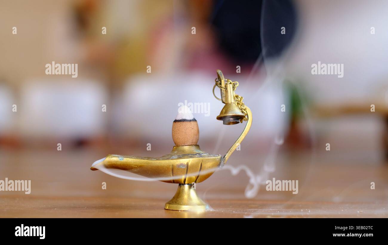 Nahaufnahme eines rauchenden Weihrauchkegels in einem Messinglampenförmigen Brenner auf einem Holzboden, mit einer meditierenden Frau, die dahinter verschwimmt. Aromatische Rituale und Stockfoto