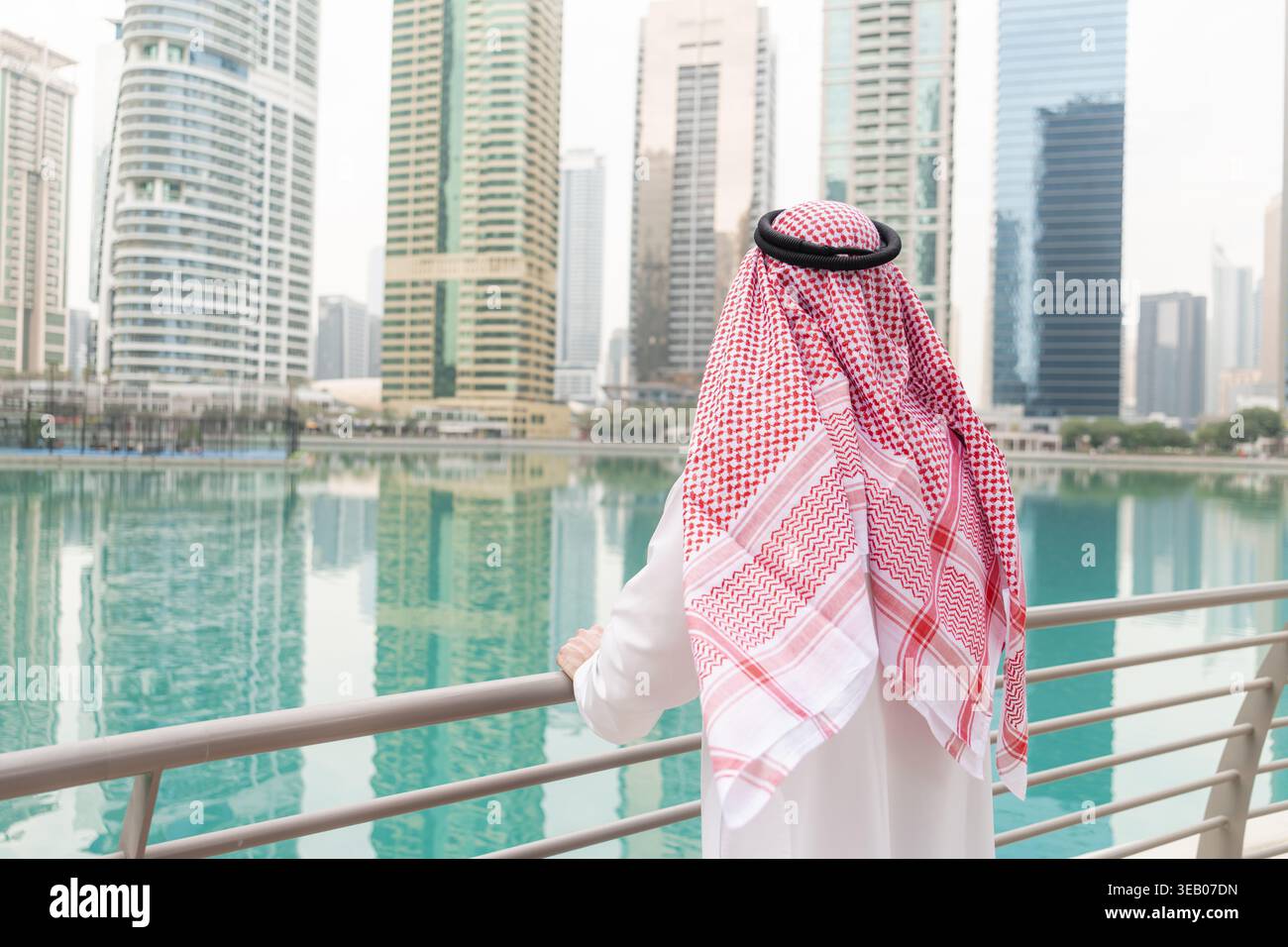 Ein arabischer Geschäftsmann steht auf einem Geländer am Wasser und blickt auf die Skyline der hohen Gebäude in einer geschäftigen Stadt. Stockfoto