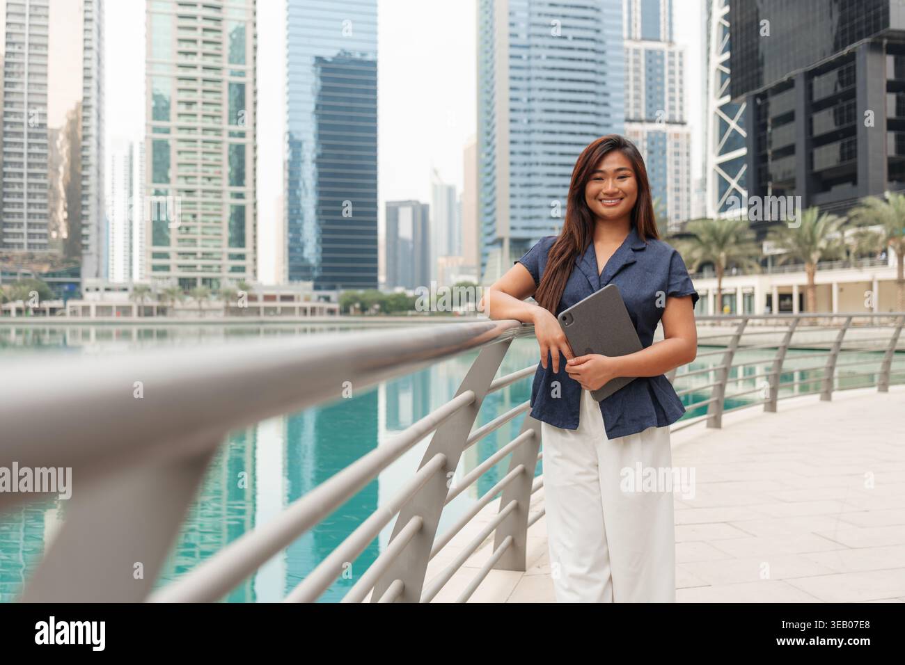 Asiatische Geschäftsfrau lächelt, während sie an klaren Tagen in einer Stadt mit hohen Gebäuden einen Laptop am Wasser hält Stockfoto