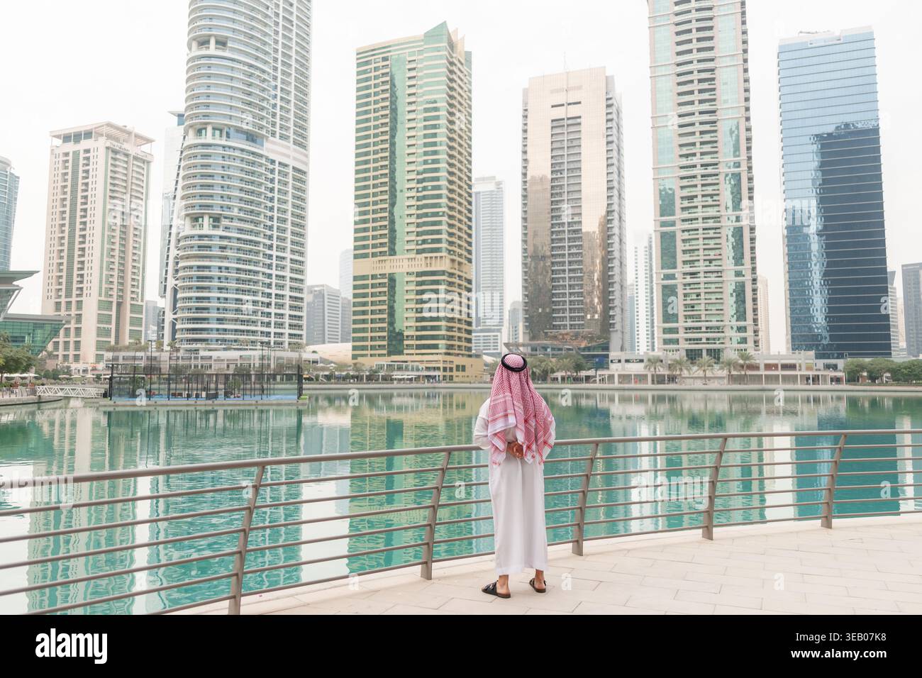 Ein arabischer Geschäftsmann steht tagsüber am Wasser und betrachtet hohe Gebäude in einer Stadt im Nahen Osten. Er trägt traditionelle Kleidung. Stockfoto