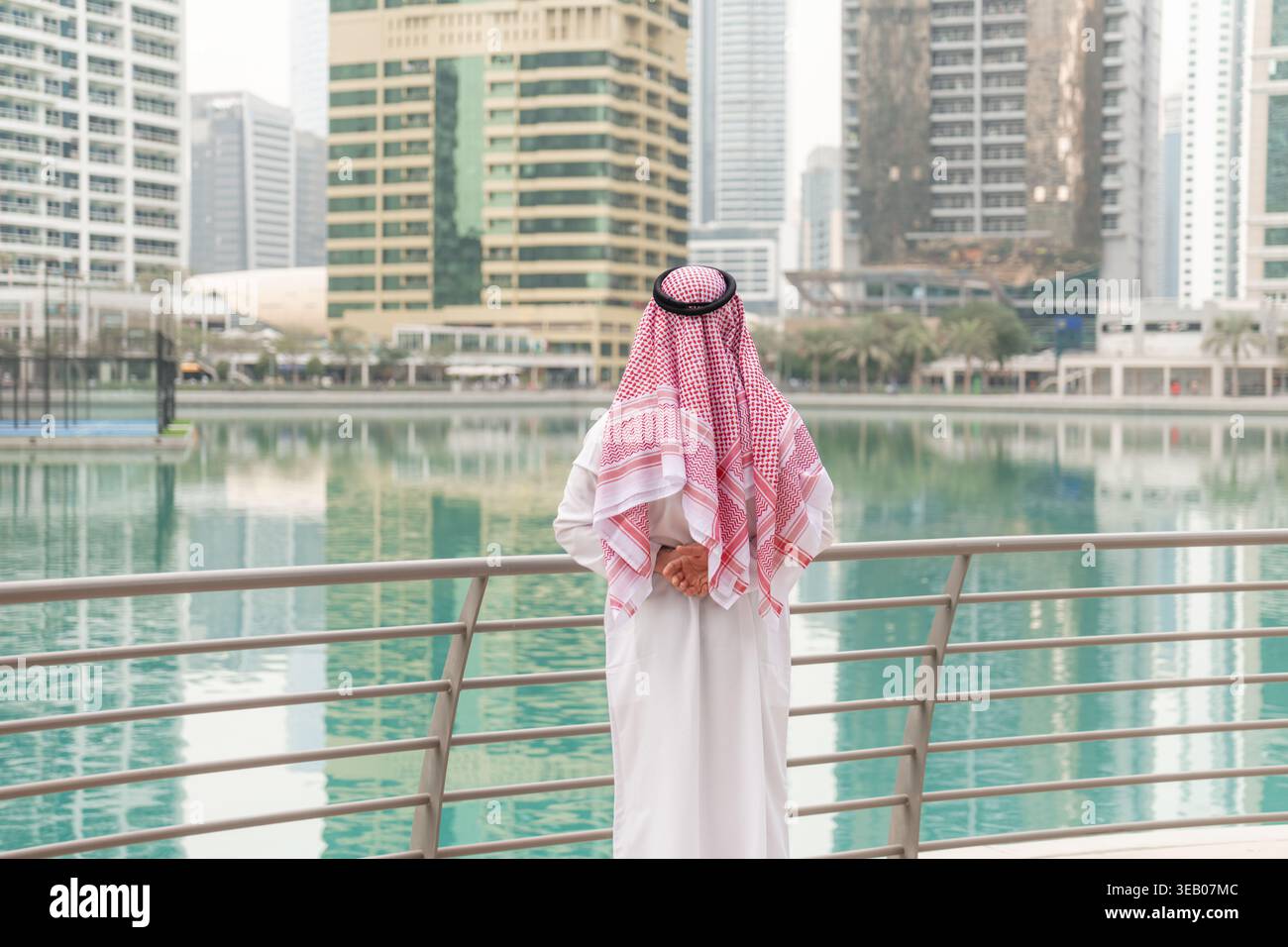 Ein arabischer Geschäftsmann steht am Wasser und blickt auf die Gebäude der Stadt im Nahen Osten. Die Sonne scheint auf die urbane Landschaft. Stockfoto