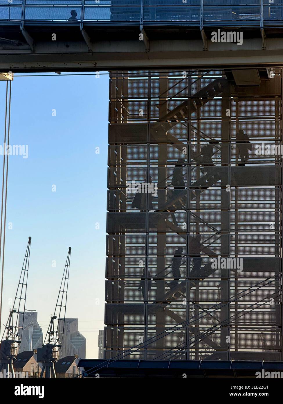 Figuren in Silhouette, die die Treppe der Royal Victoria Dock Bridge in London hinaufgehen, mit Kranichen im Hintergrund. - Smartphone-aufgenommenes Stockfoto