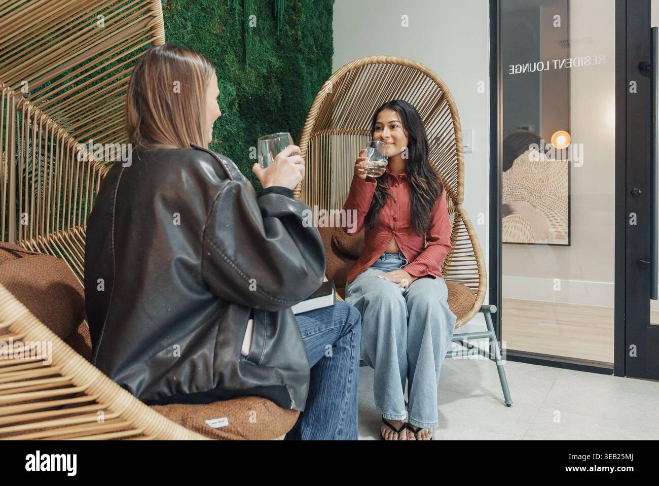 Weiße Und Asiatische Brillen Für Frauen, Die Sich In Rattan-Stühlen Mit Redaktionellem Styling, Lederjacke Und Bluse Ausgeben Stockfoto