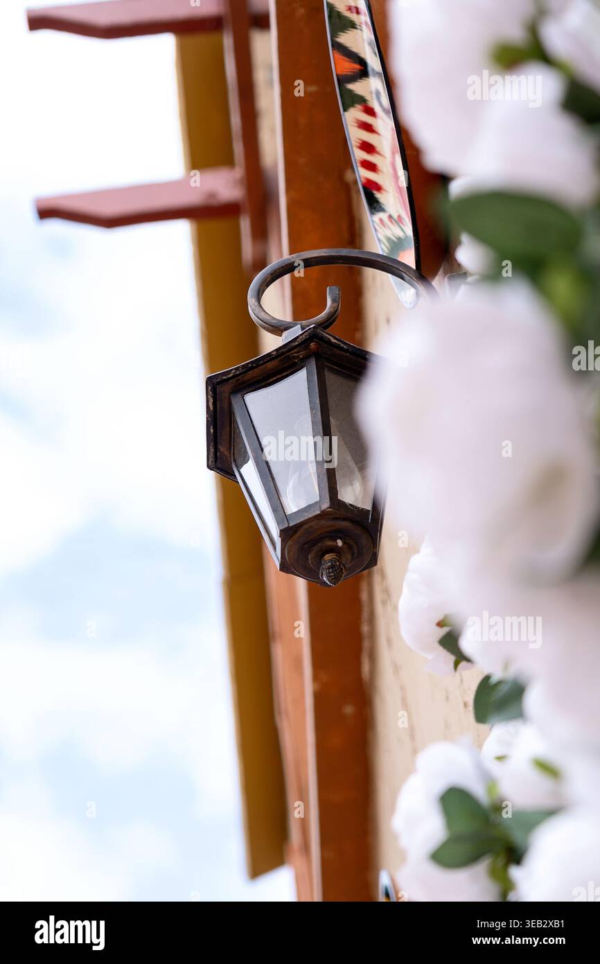 Eine alte, stilisierte hängende Laterne vor einem bewölkten blauen Himmel, bedeckt mit weißen Blumen. Das Konzept ist urbaner Komfort und Entspannung am Meer Stockfoto