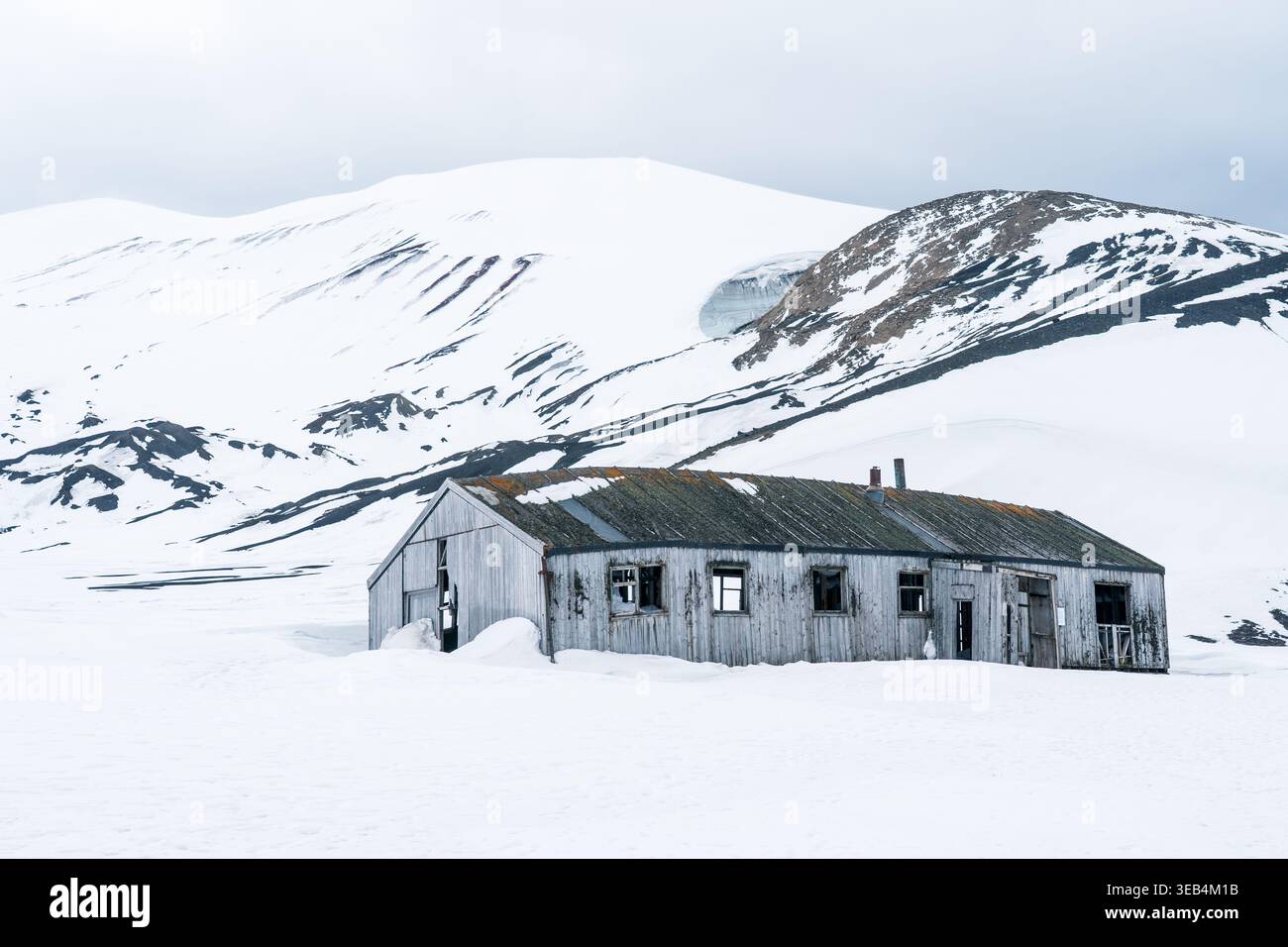 Blick auf ein verlassenes Holzgebäude auf Deception Island mit schneebedeckten Bergen und einem bewölkten Himmel in der Antarktis. Stockfoto