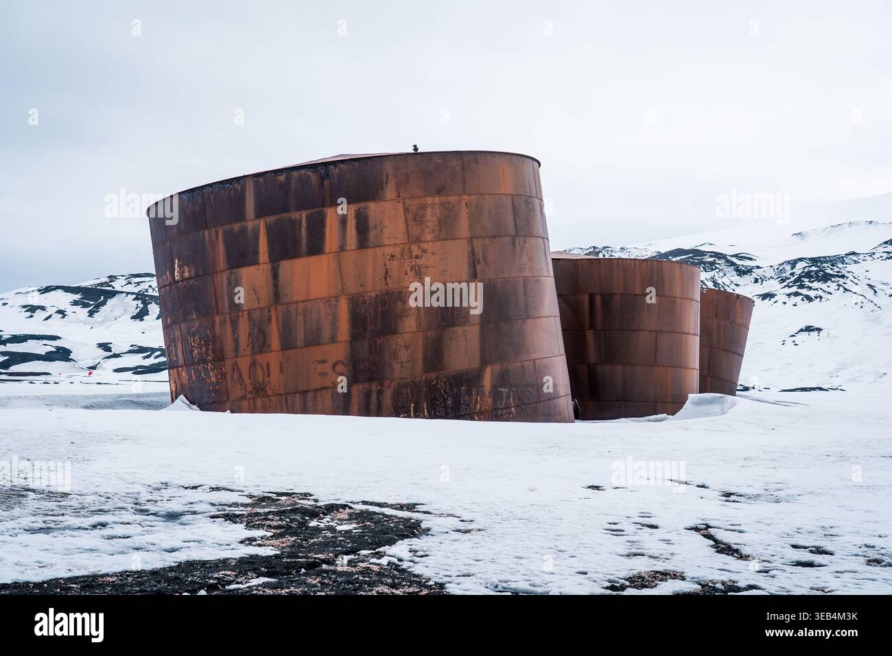 Blick auf rostige Walöltanks auf Deception Island, umgeben von schneebedeckten Bergen und vulkanischem Sand in der Antarktis. Stockfoto