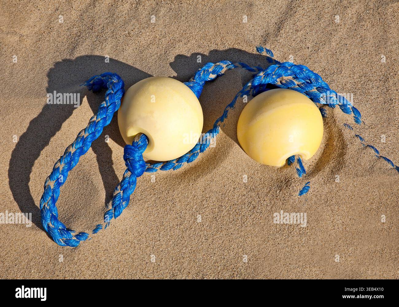 Zwei verwitterte gelbe Fischernetze, die sich in hellblauem Seil verheddern, ruhen auf sandigem Sandstrand unter warmem Sonnenlicht mit subtilen Schatten und Küstenruhe. Stockfoto