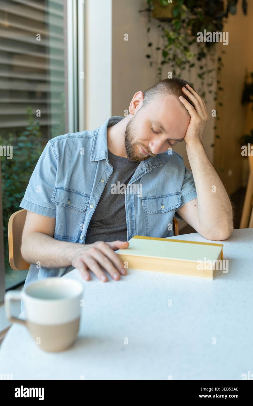 Eine Person sitzt an einem Tisch mit einem Buch vor sich. Sie haben eine Tasse Kaffee in der Nähe und sehen gestresst aus, während sie versuchen zu lernen. Das Zimmer ist gut beleuchtet Stockfoto