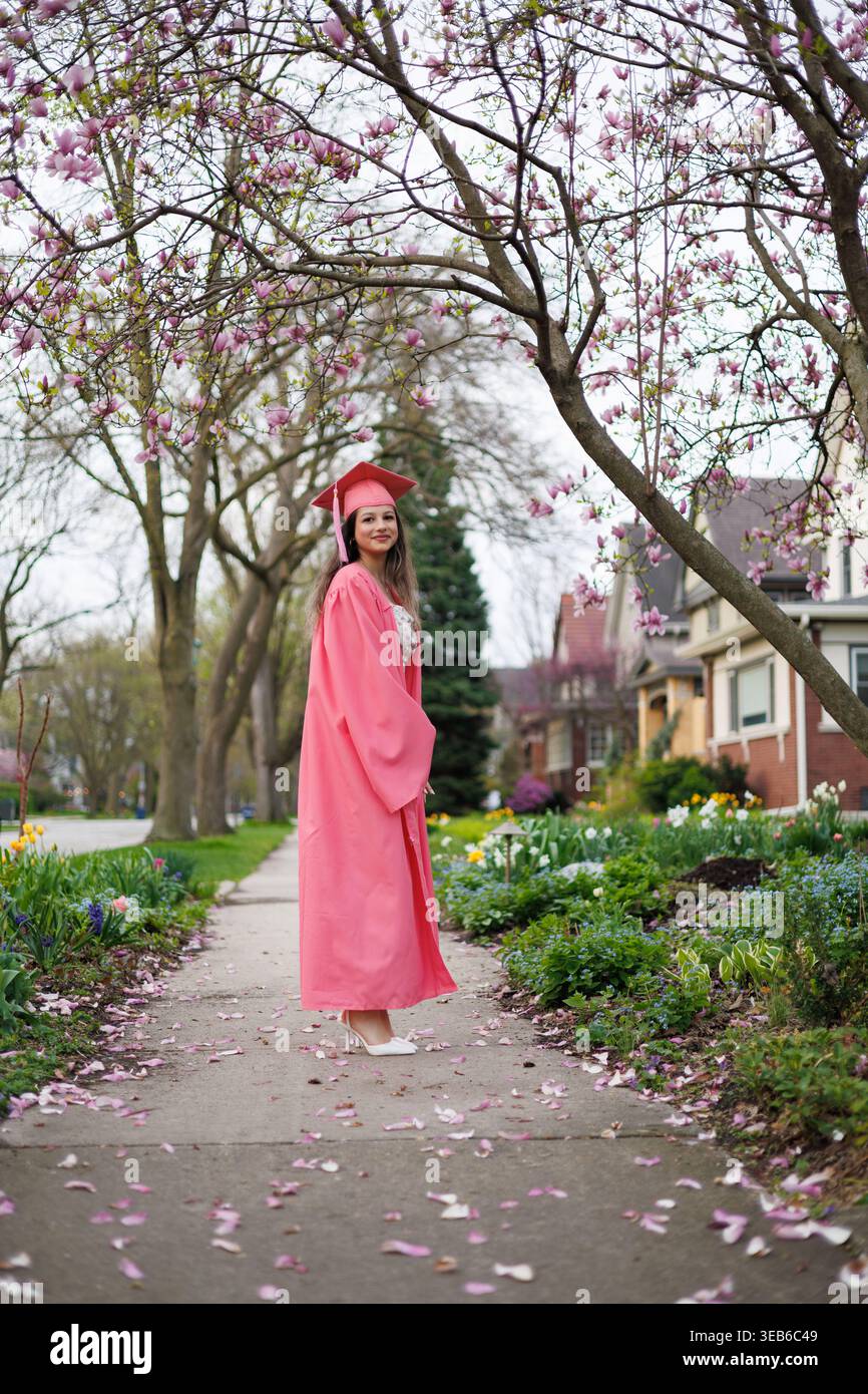Ein junger Mensch steht auf einem Bürgersteig und trägt ein rosafarbenes Abschlusskleid und eine Mütze. Blumen blühen herum und rosafarbene Blüten bedecken den Boden. Hellgrüne Bäume Stockfoto