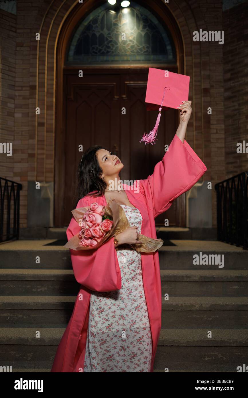 Eine Frau steht nachts auf den Stufen eines Schulgebäudes. Sie trägt ein rosafarbenes Kleid und hält ein Diplom hoch in einer Hand. In ihrer anderen Hand hat sie das Stockfoto