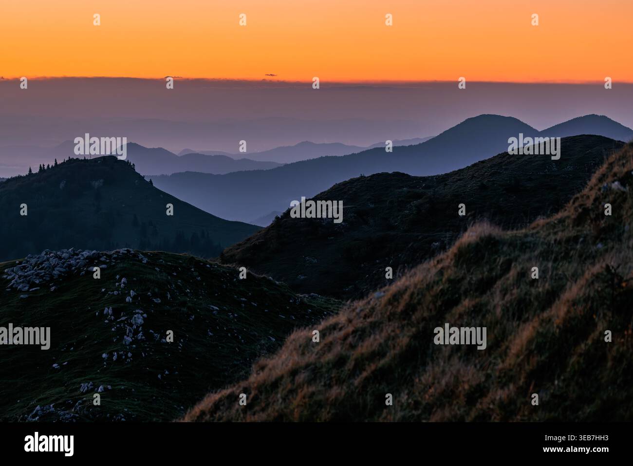 Blick auf mehrschichtige Bergsilhouetten unter einem leuchtend orangefarbenen Sonnenuntergangshimmel mit nebeligen Tälern in Sorica, Skofja Loka, Slowenien. Stockfoto