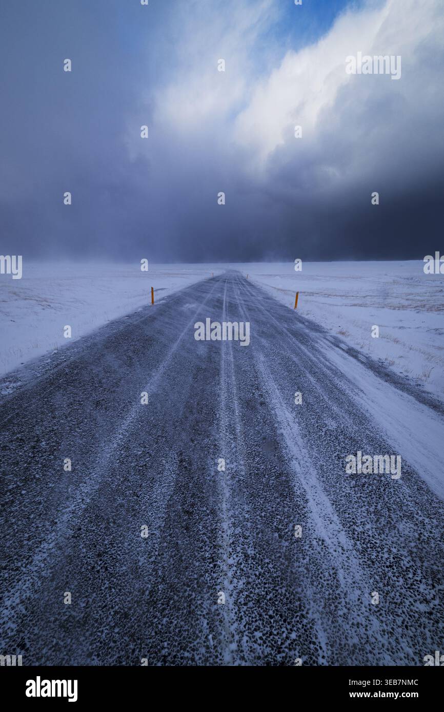Blick auf eine gerade, mit Schnee und Eis bedeckte Asphaltstraße, die sich durch eine weite Winterlandschaft unter einem dramatischen bewölkten Himmel in Snaefellsnes, Eyja- Stockfoto