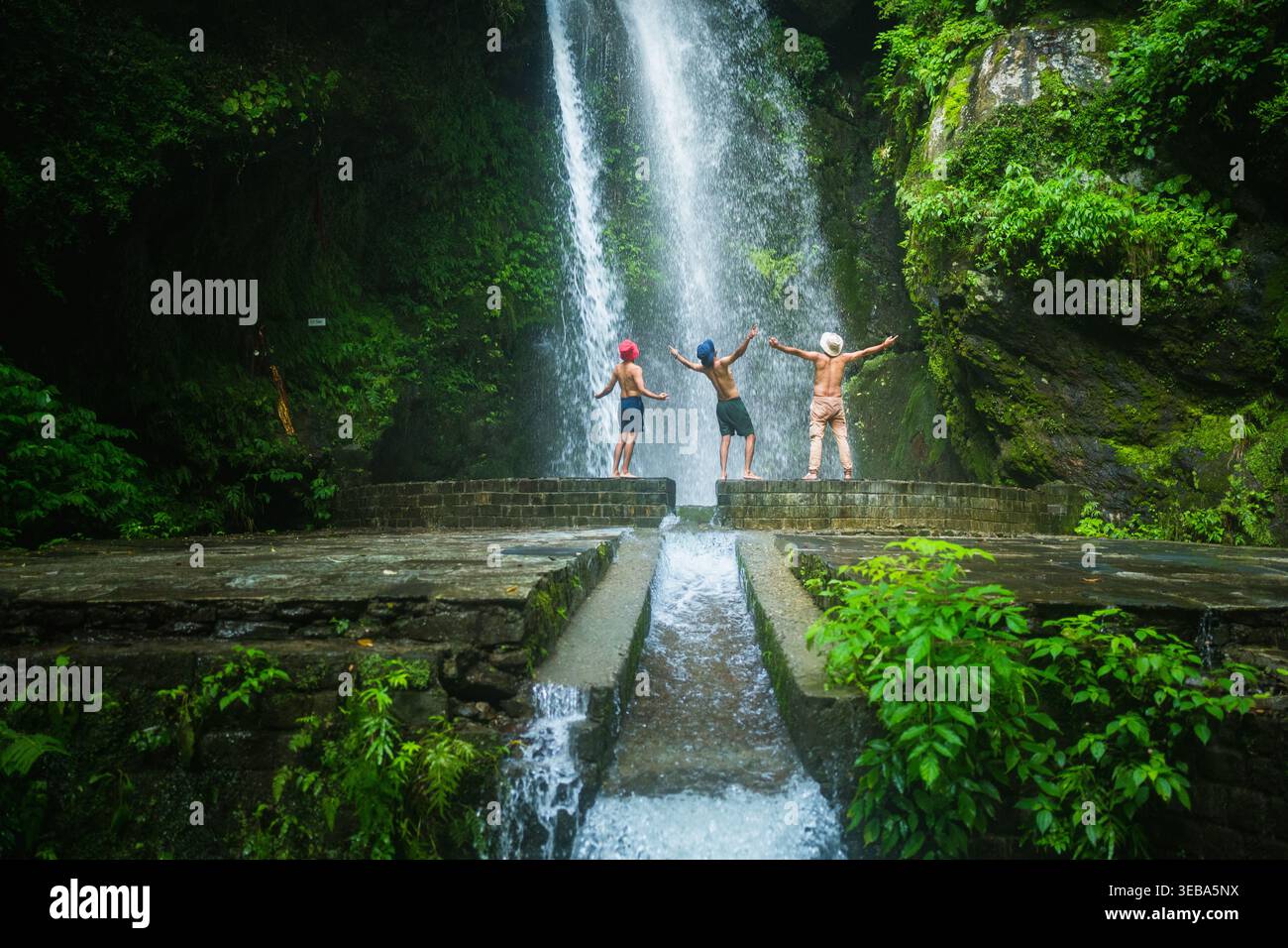 Drei Freunde stehen mit ausgebreiteten Armen vor einem mächtigen Wasserfall und schwelgen in der atemberaubenden Naturschönheit und ihrer gemeinsamen abenteuerlichen Reise i Stockfoto