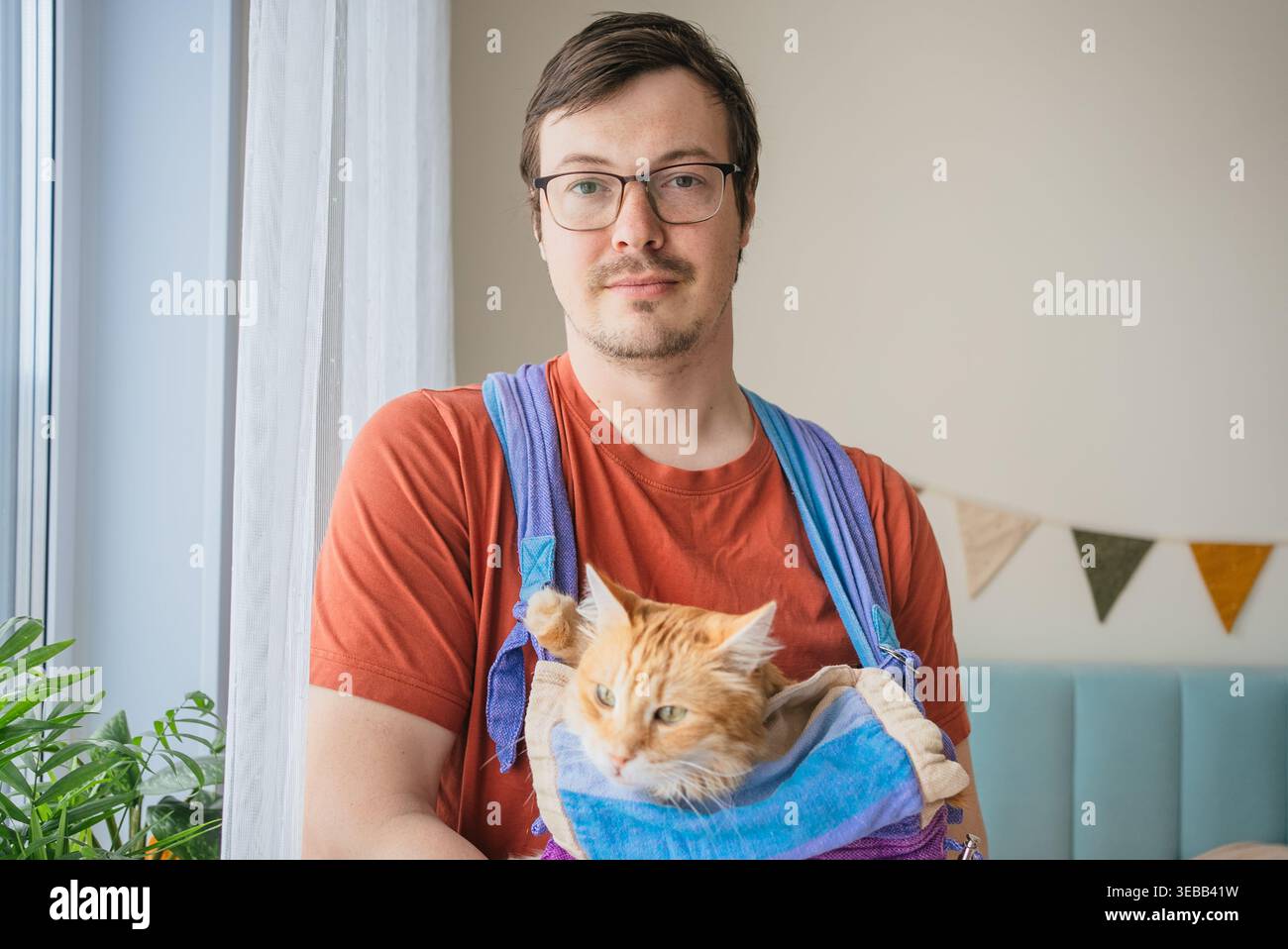 Ein Mann, der eine Ingwerkatze trägt, schmiegt sich bequem in eine lebendige Haustierschlinge, verkörpert Tierbesitz, Tierkomfort und die einzigartige Verbindung zwischen Menschen und ihren katzenartigen Begleitern im Haus Stockfoto