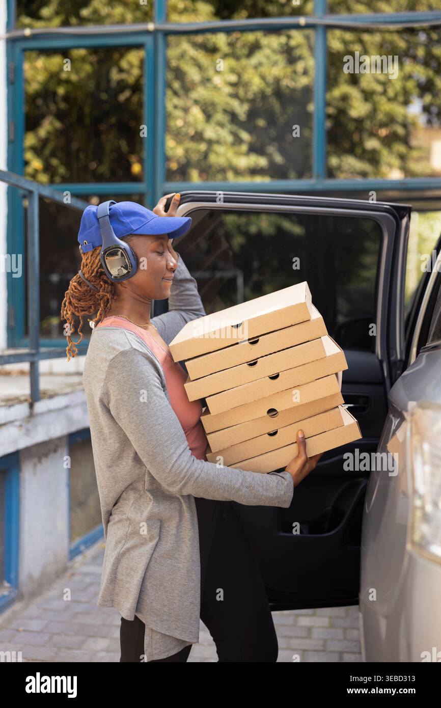 Schwarzer Mitarbeiter zum Mitnehmen parkt das Auto, um Pizzakartons vor der Haustür zu bringen und eine riesige Fast-Food-Bestellung zu liefern. Weiblicher Kurierdienst, der einen 5-Sterne-Express-Service sicherstellt. Stockfoto