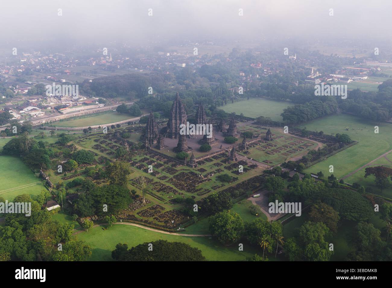Blick aus der Vogelperspektive auf den Prambanan Tempel, den alten hinduistischen Tempelkomplex mit hoch aufragenden Steintürmen, umgeben von grünen Rasenflächen und nebligem Dunst in Kecamatan Pra Stockfoto