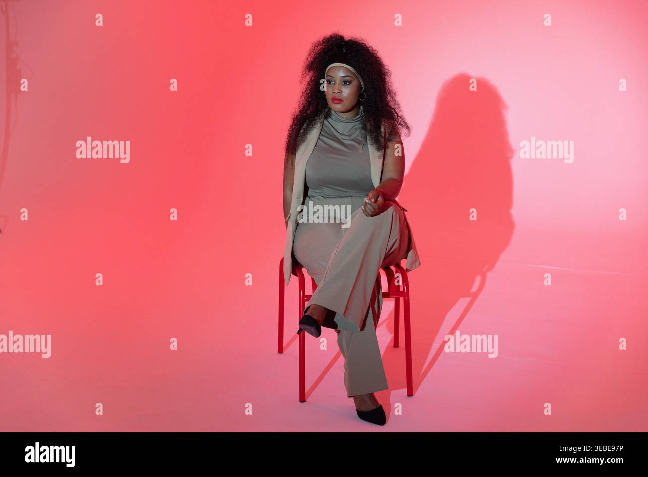 Afroamerikanerin im mittleren Erwachsenenalter sitzt auf rotem Metallstuhl, trägt neutrales Outfit und wirft Schatten Stockfoto