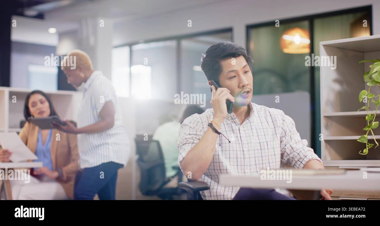 Sprechender asiatischer Mann, der ein leichtes kariertes Hemd mit gerollten Ärmeln im offenen Büro trägt, am Telefon Stockfoto