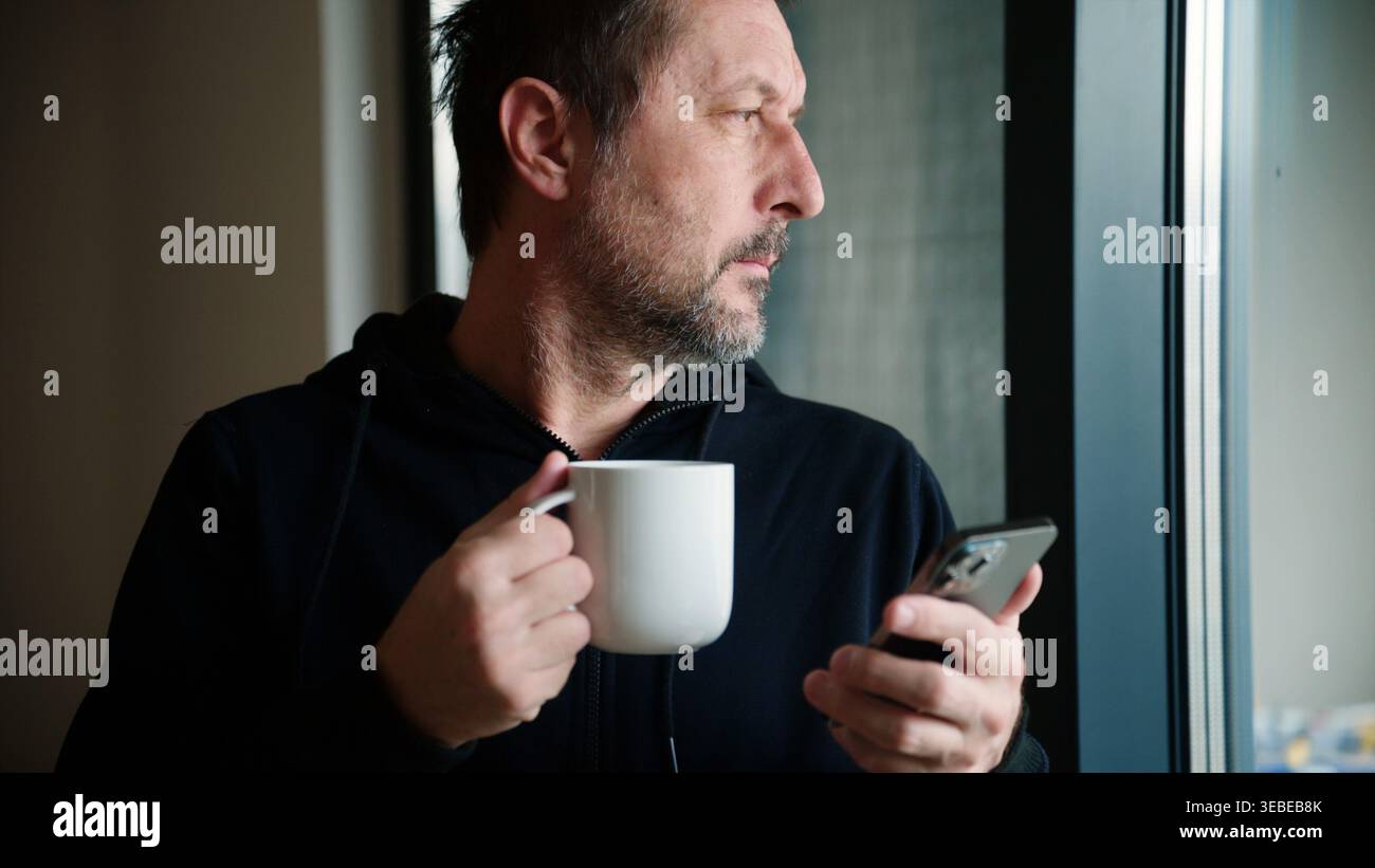 Mann mittleren Alters, der in sozialen Medien am Telefon surft, während er Kaffee hält, im ruhigen Morgenmoment aus dem Fenster blickt, selektiver Fokus Stockfoto
