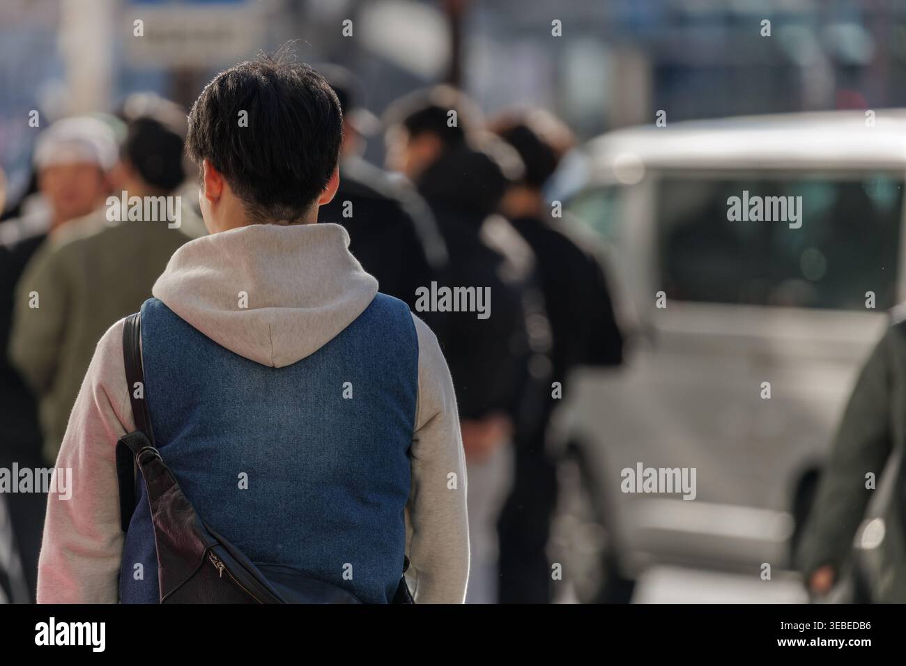 Junger Mann in mehrschichtiger Streetwear aus Jeansweste und Hoodie, der in der Menge auf der belebten Stadtstraße spaziert. Die offene Szene fängt Anonymität und den Alltag ein. Stockfoto