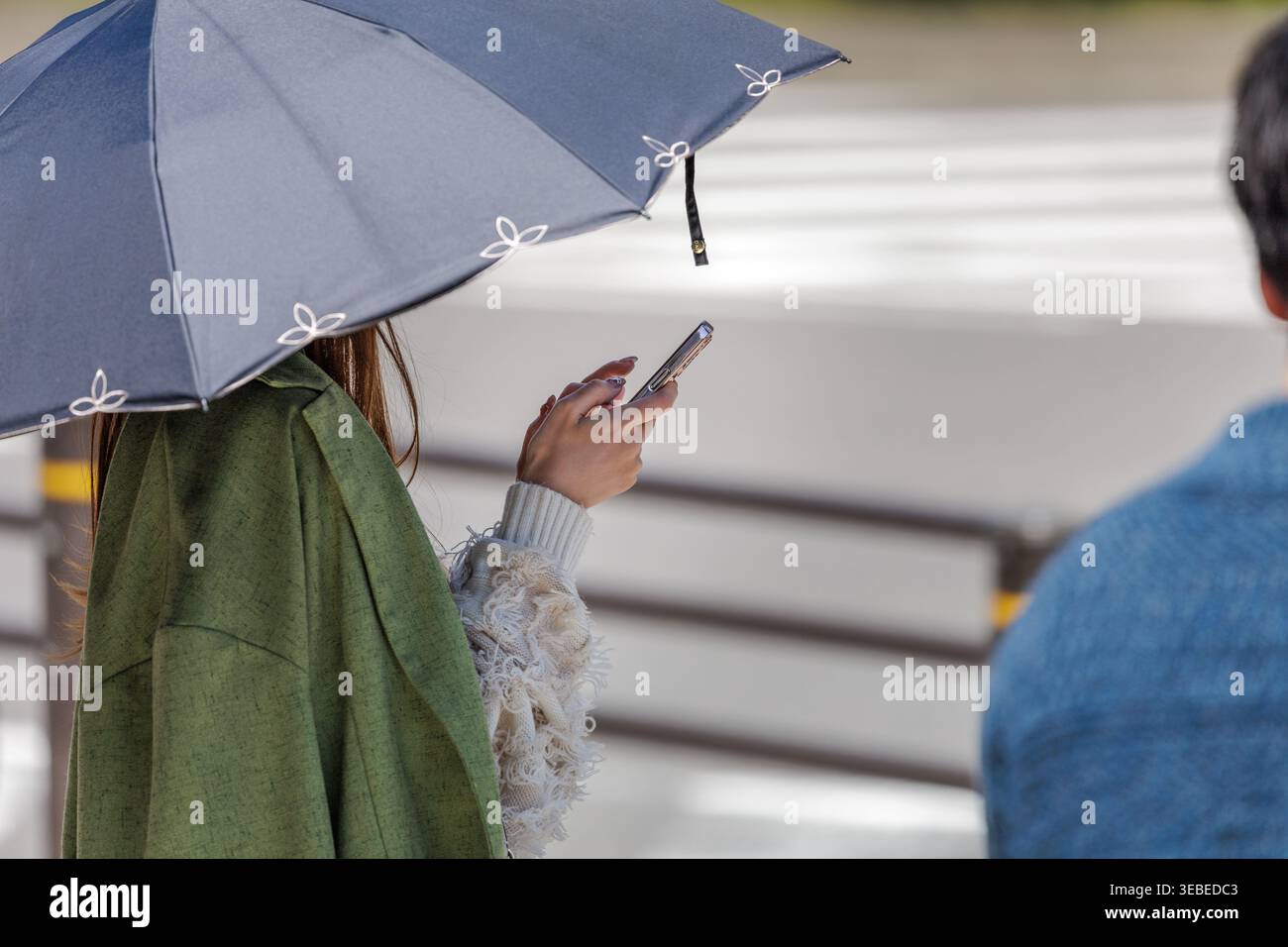 Mantel über der Schulter drapiert, schicker Schirm in der Hand, alltägliche Eleganz zeigt sich, während die Frau auf den Crosswalk wartet und das Telefon mit ihrer freien Hand leicht gehalten wird. Stockfoto