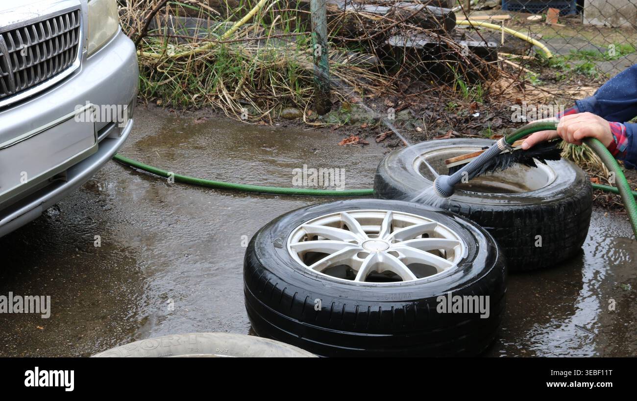 Waschen und Reinigen von Autorädern mit einem Wasserstrahl aus einem Schlauch mit einer Gießkanne, die auf dem Asphalt neben dem Auto liegt und Räder pflegt Stockfoto