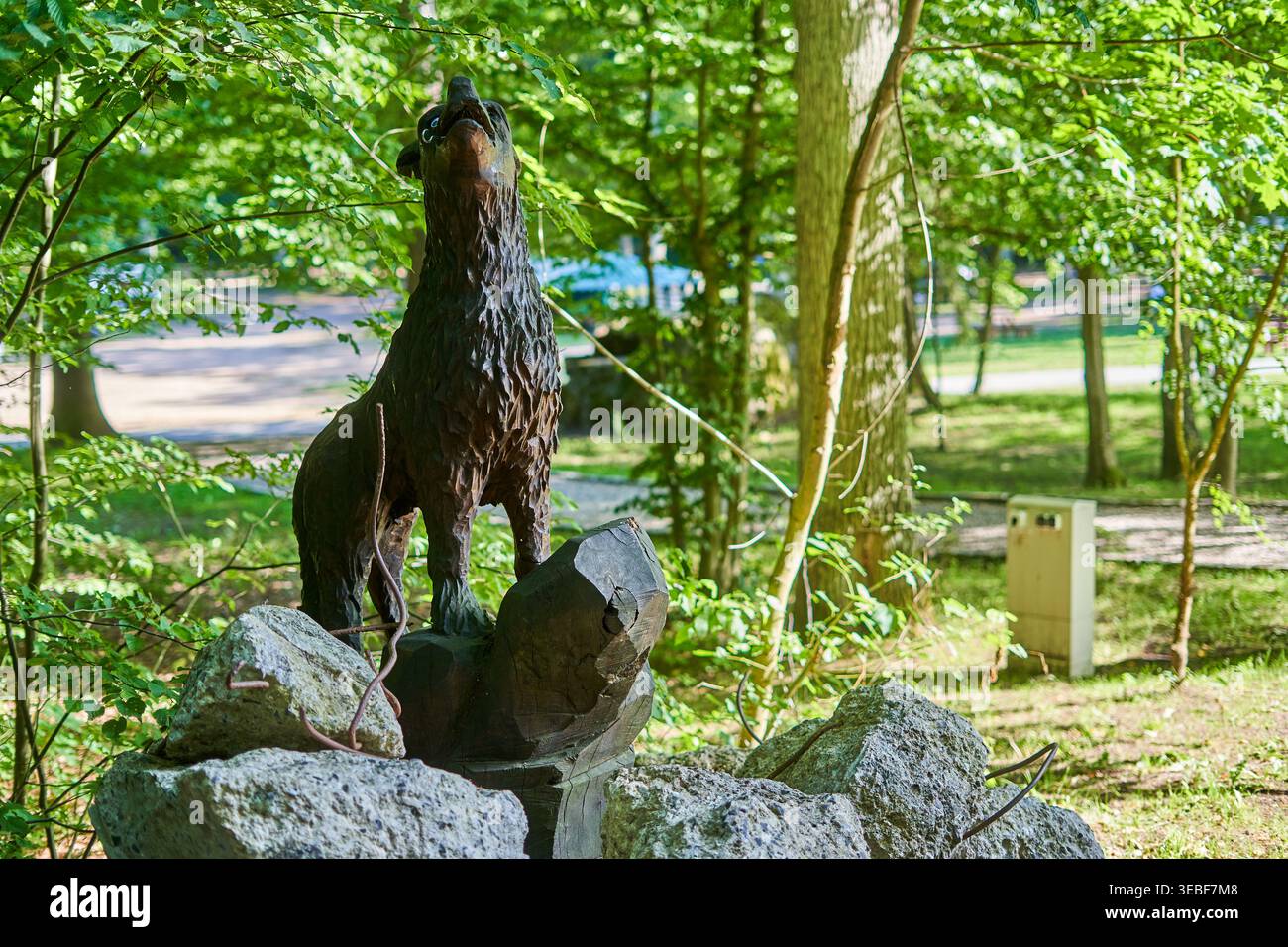 Eine Bronzestatue eines heulenden Wolfes steht auffällig auf einem felsigen Steinpodest in einem üppigen grünen Waldpark. Die Skulptur fängt das Tier mit Stockfoto