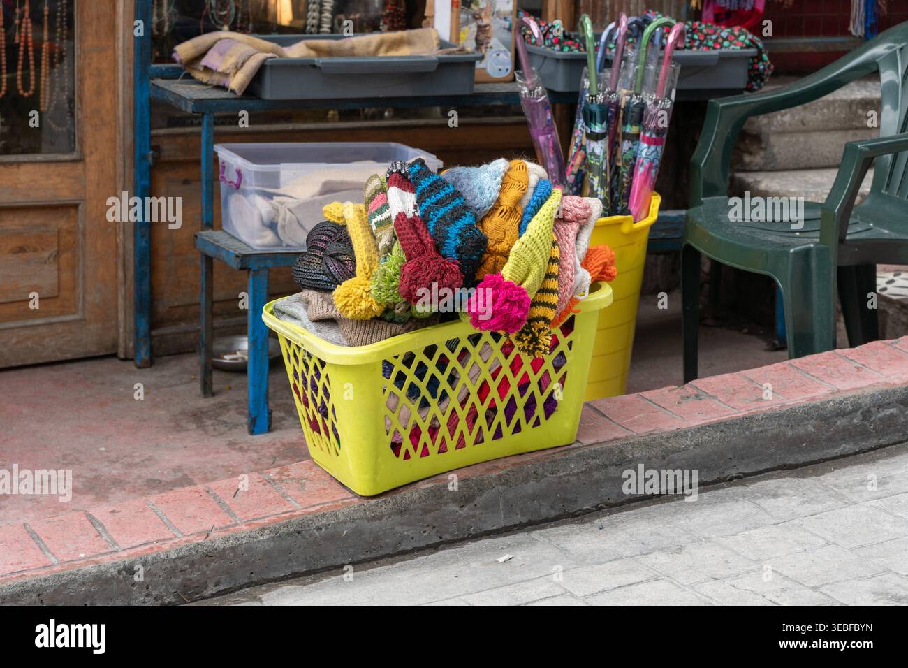 Bunte Strickmützen und Tücher in einem Plastikkorb in der Nähe des Eingangs zum Straßenladen mit Regenschirmen und Ausstellungsartikeln. Einzelhandelsszene, Straßenmarkt, Mode A Stockfoto